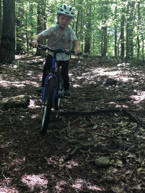 A child riding a blue bicycle on a wooded dirt trail, surrounded by green trees and sunlight filtering through the leaves. The child is wearing a white helmet and is focused on navigating the terrain. The ground is covered with rocks and fallen leaves, illustrating the natural environment. Green River mountain bike trail.