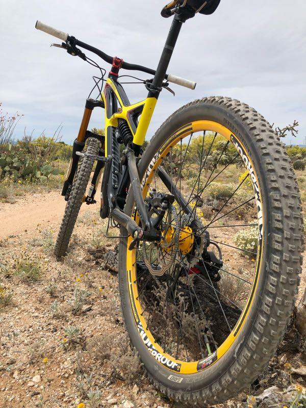 A close-up view of a mountain bike resting on a dirt trail, with its yellow and black frame prominently displayed. The bike's rear wheel is in focus, showcasing its rugged tire design, surrounded by desert vegetation and a cloudy sky in the background. Fantasy Island mountain bike trail.