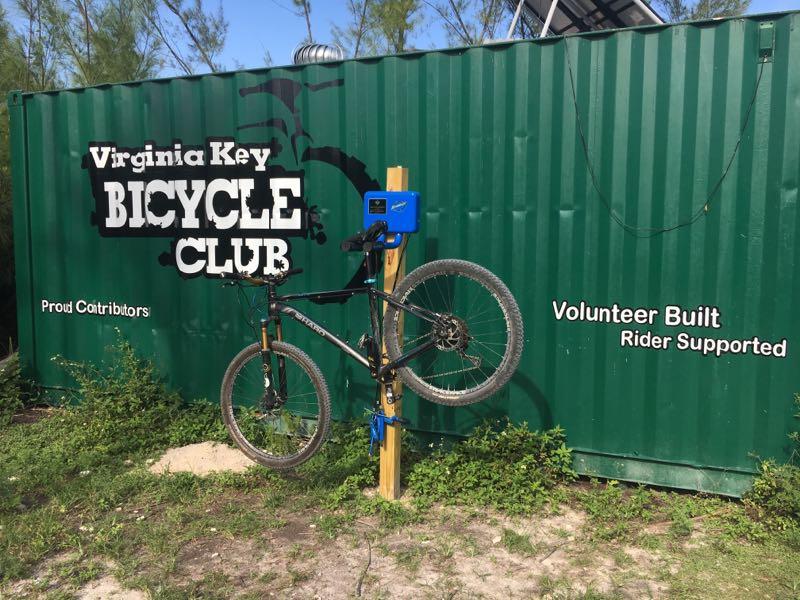 A black mountain bike suspended on a repair stand in front of a green shipping container featuring the text "Virginia Key BICYCLE CLUB" and "Volunteer Built Rider Supported." The scene is set in a grassy area with some surrounding foliage. Virginia Key North Point mountain bike trail.