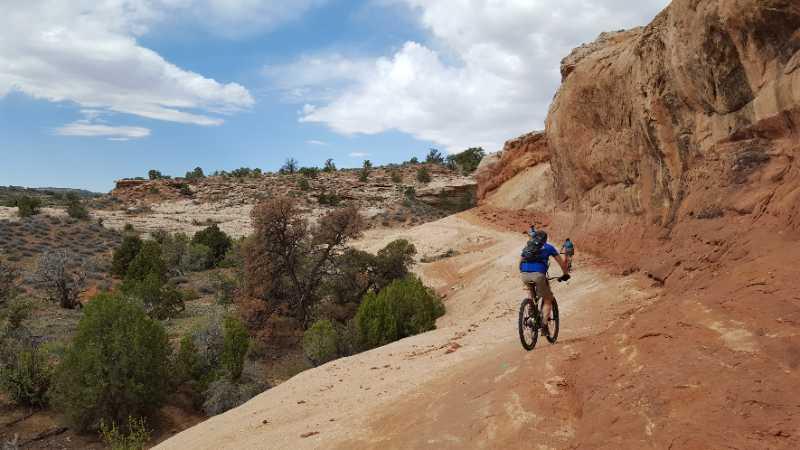 Two mountain bikers ride along a rugged dirt trail in a desert landscape characterized by rocky formations and sparse vegetation. The sky is partly cloudy, creating a vivid backdrop for their adventure. Rocky Tops mountain bike trail.