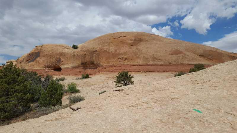 A rocky landscape featuring a large, gently sloping hill with reddish-brown layers and sparse vegetation, including small trees and shrubs. The sky above is partly cloudy, with scattered clouds. The terrain is dry and rocky, characteristic of a desert environment. Rocky Tops mountain bike trail.