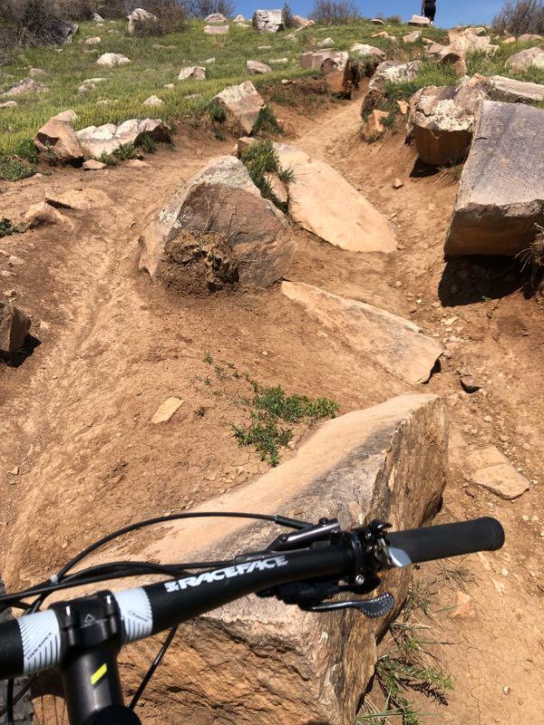 Mountain bike handlebars are visible in the foreground, pointing toward a rocky trail with loose dirt and grass. Large boulders line the path, which winds up a grassy slope under a clear blue sky. A person can be seen in the background, walking along the trail. Foothills Trail mountain bike trail.