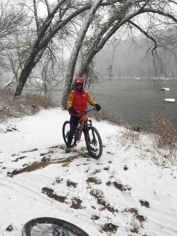 A person in a bright red vest and orange sleeves is standing next to a mountain bike on a snow-covered path by a river, with trees in the background and snow falling gently. Rappahanock River Trail mountain bike trail.