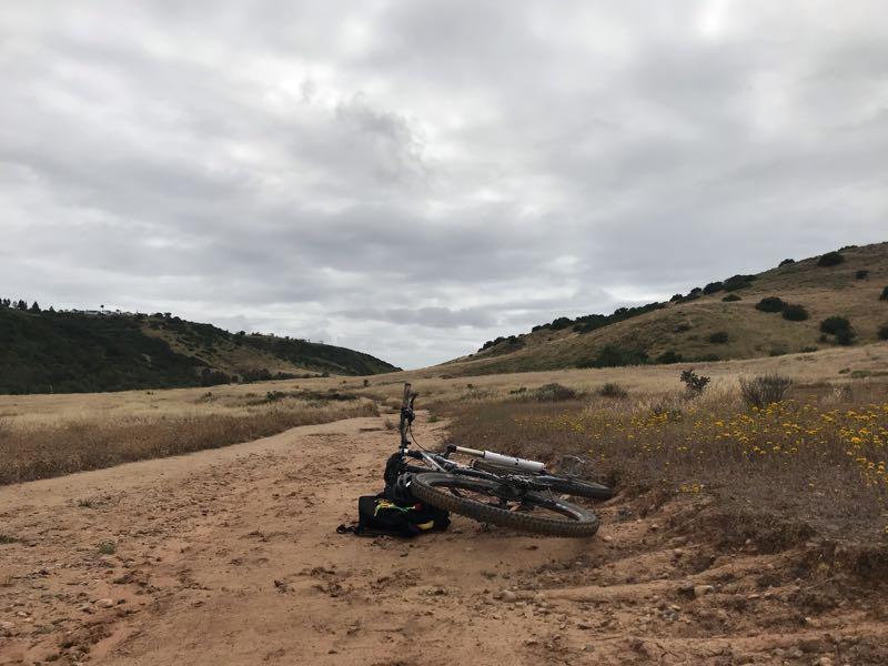 A mountain bike lies on its side on a dirt path, surrounded by open fields and rolling hills under a cloudy sky. The landscape features patches of wildflowers and sparse vegetation, creating a serene outdoor scene. Los Penasquitos Canyon Preserve mountain bike trail.