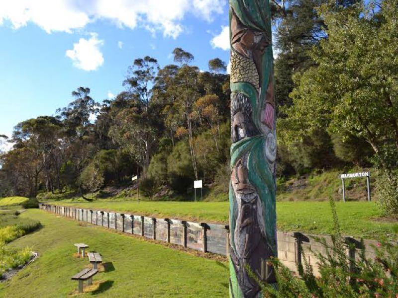 A scenic view of a park featuring a prominently displayed carved wooden totem pole, adorned with intricate designs representing nature and culture. In the background, there are trees and signs indicating the location, with benches along a pathway. The sky is partly cloudy, adding to the tranquil atmosphere of the setting. Lilydale to Warburton Rail Trail mountain bike trail.