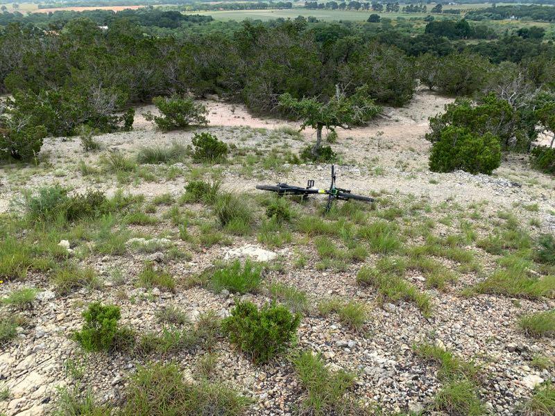 Mountain bike lying on rocky terrain with grass and scattered shrubs, overlooking a green landscape in the background. Fredericksburg Hilltop Trail mountain bike trail.