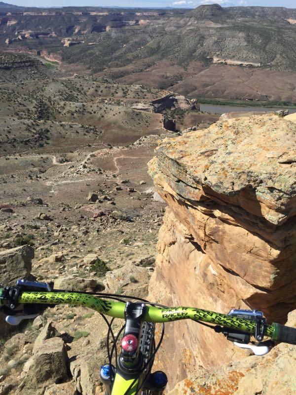 Aerial view from a mountain bike handlebar, overlooking a rocky landscape and valley. The scene features rugged terrain, cliffs, and a winding river in the distance under a clear blue sky. Kokopelli Trail mountain bike trail.