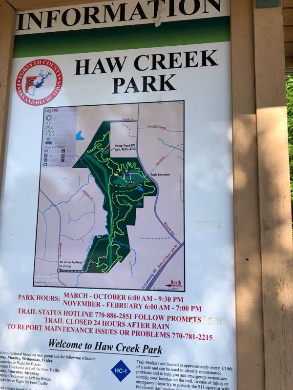 A sign displaying information about Haw Creek Park, including a map of the park's trails and facilities, park hours, and contact information for maintenance issues. The sign is prominently labeled “HAW CREEK PARK” and includes details about park hours and a hotline for trail status updates. Haw Creek Park mountain bike trail.