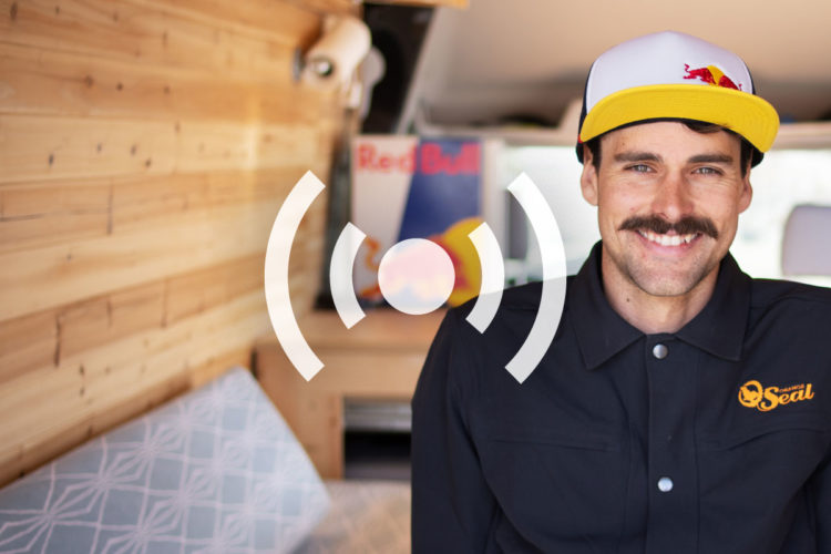 A smiling man with a thick mustache wearing a white and yellow cap sits in a stylish camper van. The background features wooden walls and soft furnishings, creating a cozy atmosphere. A logo is visible on his shirt, and there are colorful posters in the van.