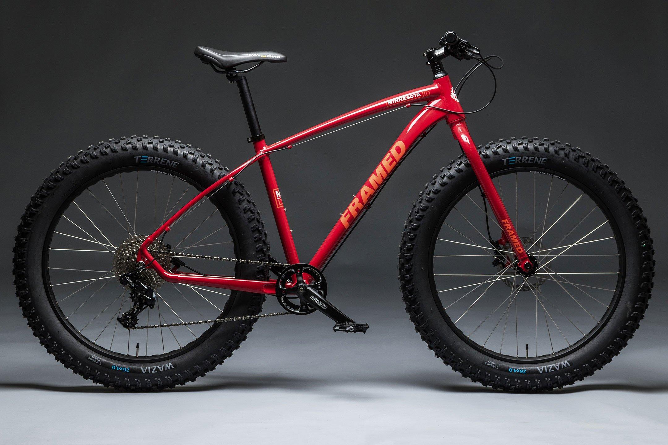 A red fat tire mountain bike positioned against a dark background, showcasing its sturdy frame, wide tires, and detailed components. The bike features black wheels with prominent tire treads and a visible drivetrain.