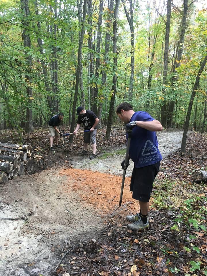 Three individuals are working together in a wooded area, raking and leveling soil along a pathway. They appear focused on their task, surrounded by trees with green and yellow foliage. In the background, there are logs stacked, indicating recent clearing work. The ground is damp and covered with fallen leaves, typical of a forested environment. Angler's Ridge mountain bike trail.