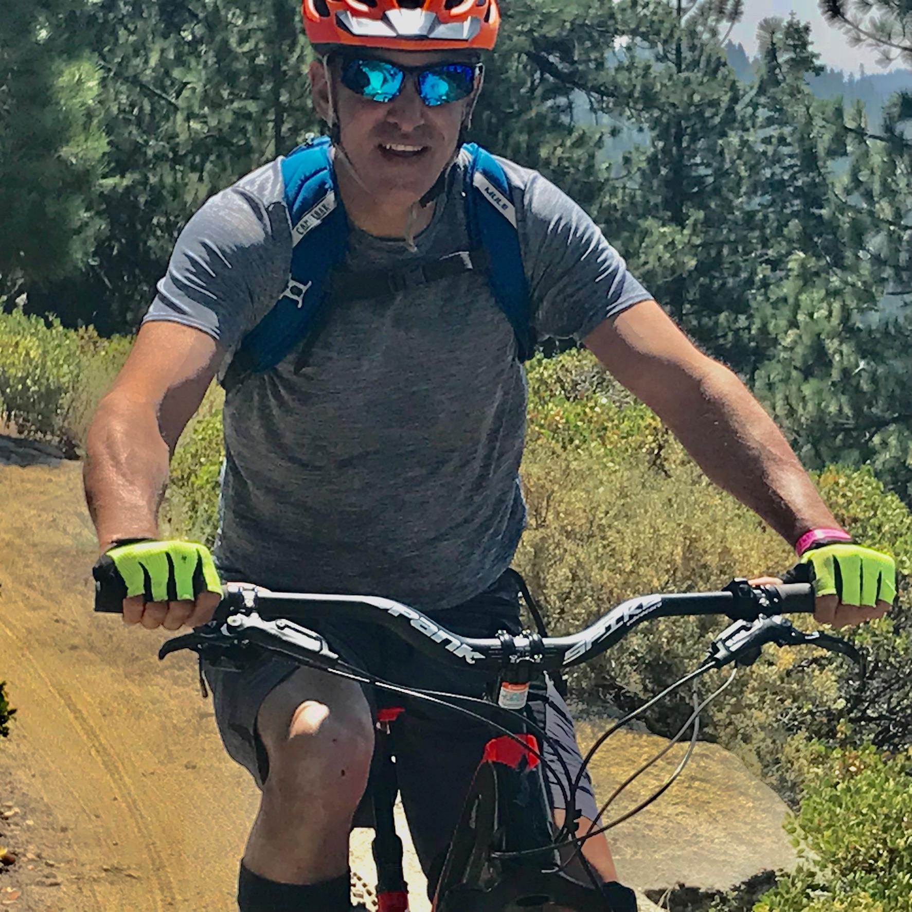 Specialized Levo SFR Expert 6Fattie: A person riding a mountain bike on a dirt trail surrounded by greenery. They are wearing a grey athletic shirt, orange helmet, sunglasses, and colorful gloves. A blue backpack is visible on their back as they pedal uphill.
