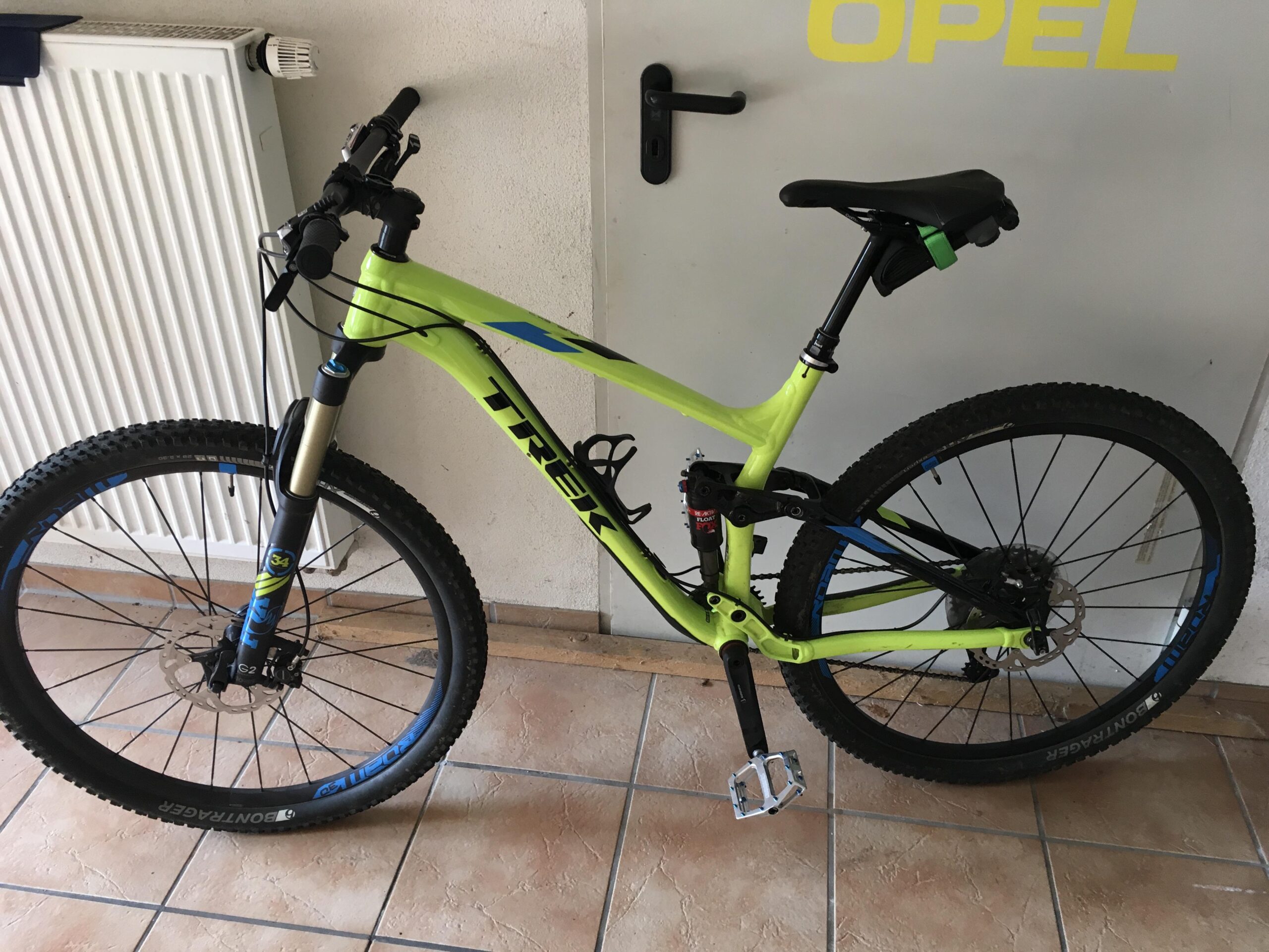 Trek Fuel EX 9 29: A bright yellow Trek mountain bike positioned indoors, showcasing its front wheel and handlebars. The bike features thick tires designed for off-road use, a dual suspension system, and a sleek frame. In the background, there is a white radiator and a door with a yellow "OPEL" logo. The flooring consists of light-colored tiles.