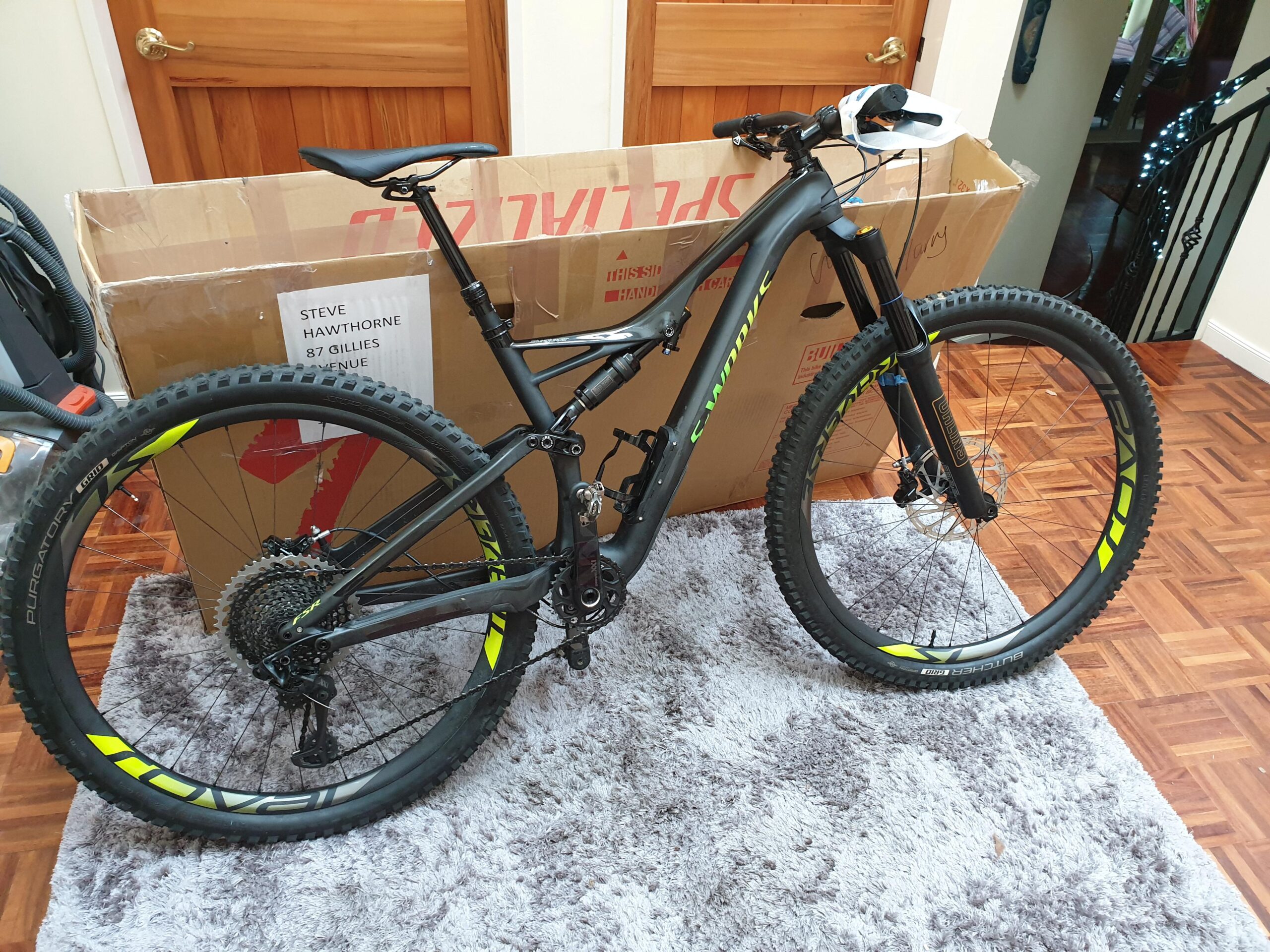 Specialized SWORKS Stumpjumper FSR: A new black mountain bike with green accents is partially assembled, resting on a plush rug. In the background, a large cardboard box is visible, along with a label featuring a name and address. The setting includes wooden doors and a staircase.