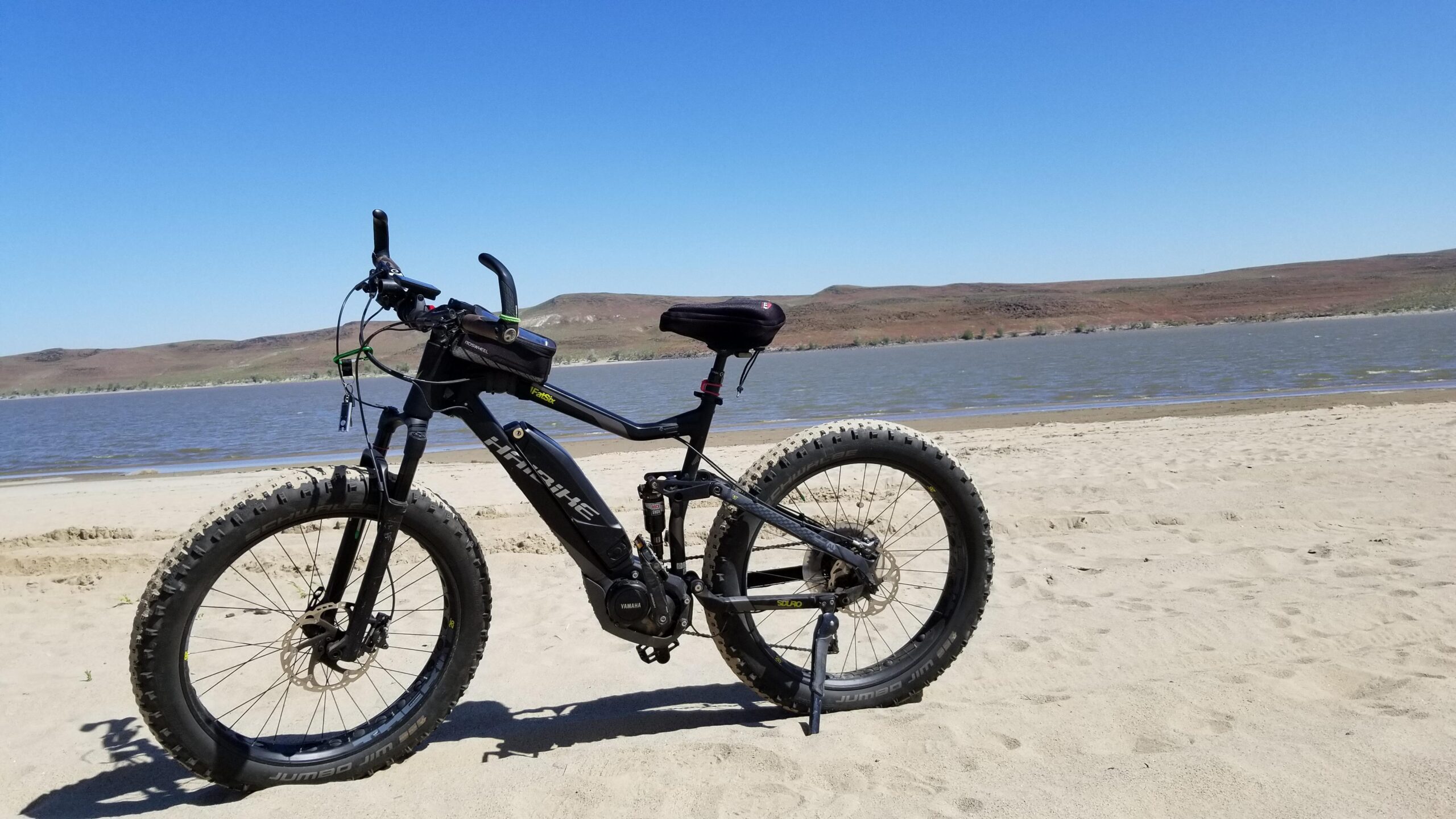Haibike Fat Six: An electric fat bike on a sandy beach, with a body of water and hills in the background under a clear blue sky. The bike features thick tires, a comfortable seat, and a small bag mounted on the frame.