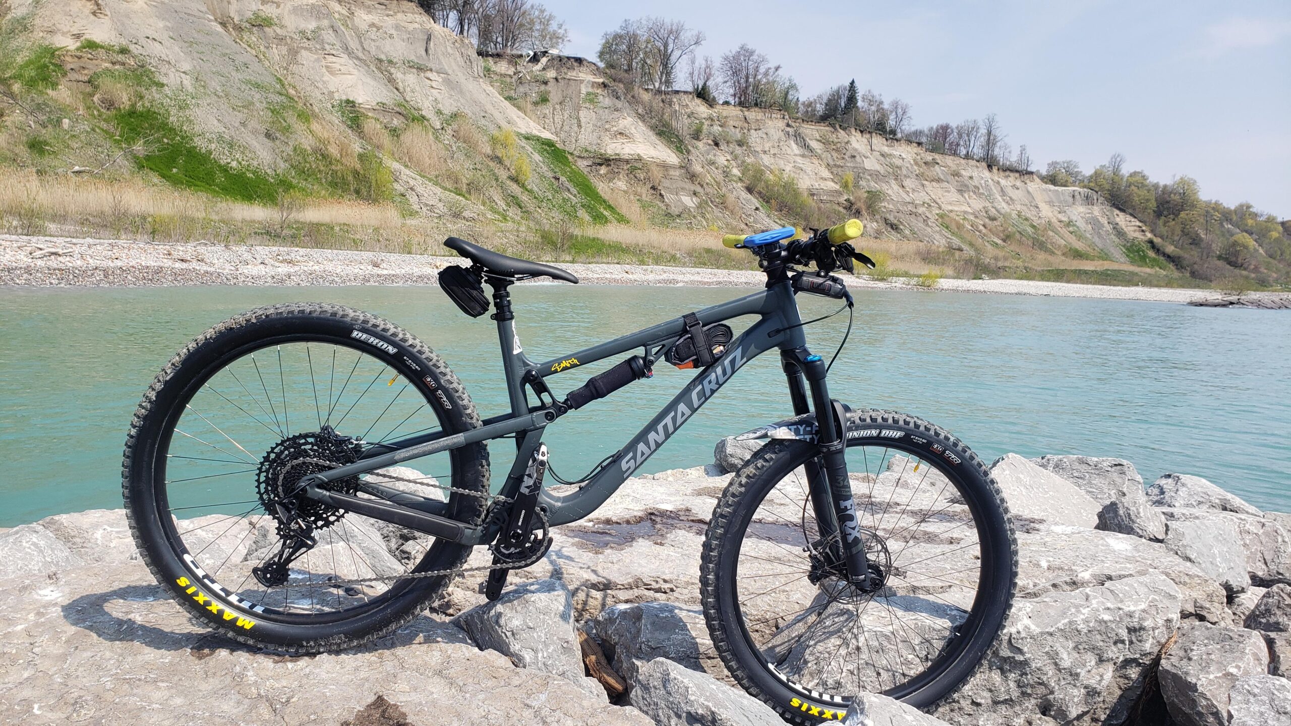 Santa Cruz 5010: A mountain bike is positioned on large rocks near a body of water, with a background of grassy hills and a clear blue sky. The bike features a gray frame with yellow accents on the tires and is equipped for off-road riding.