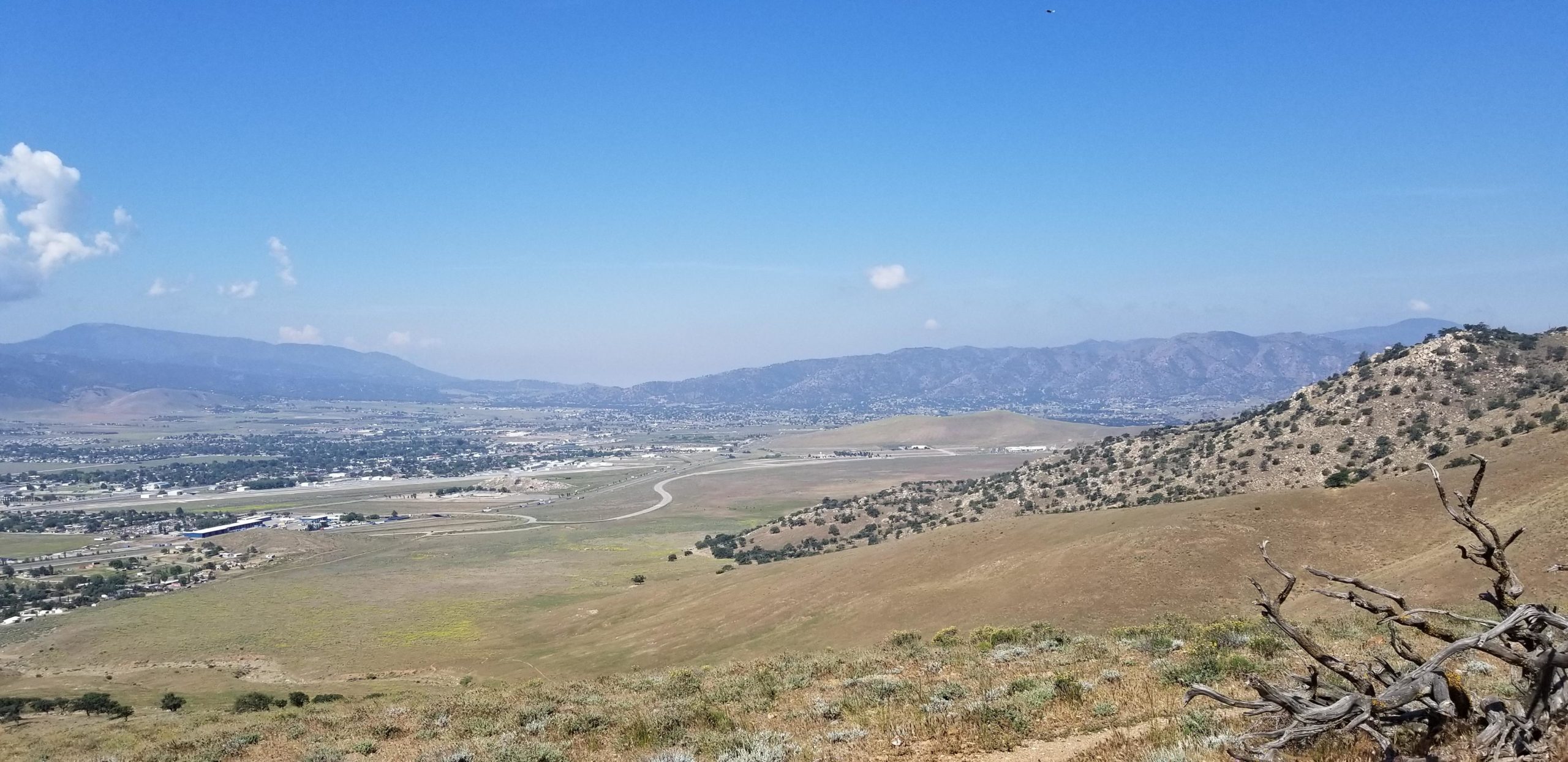 A panoramic view of rolling hills and valleys under a clear blue sky, featuring distant mountains on the horizon. The landscape includes patches of green vegetation and dry terrain, with winding roads visible in the foreground and small buildings scattered throughout the valley below. TMTA Lehigh trails mountain bike trail.