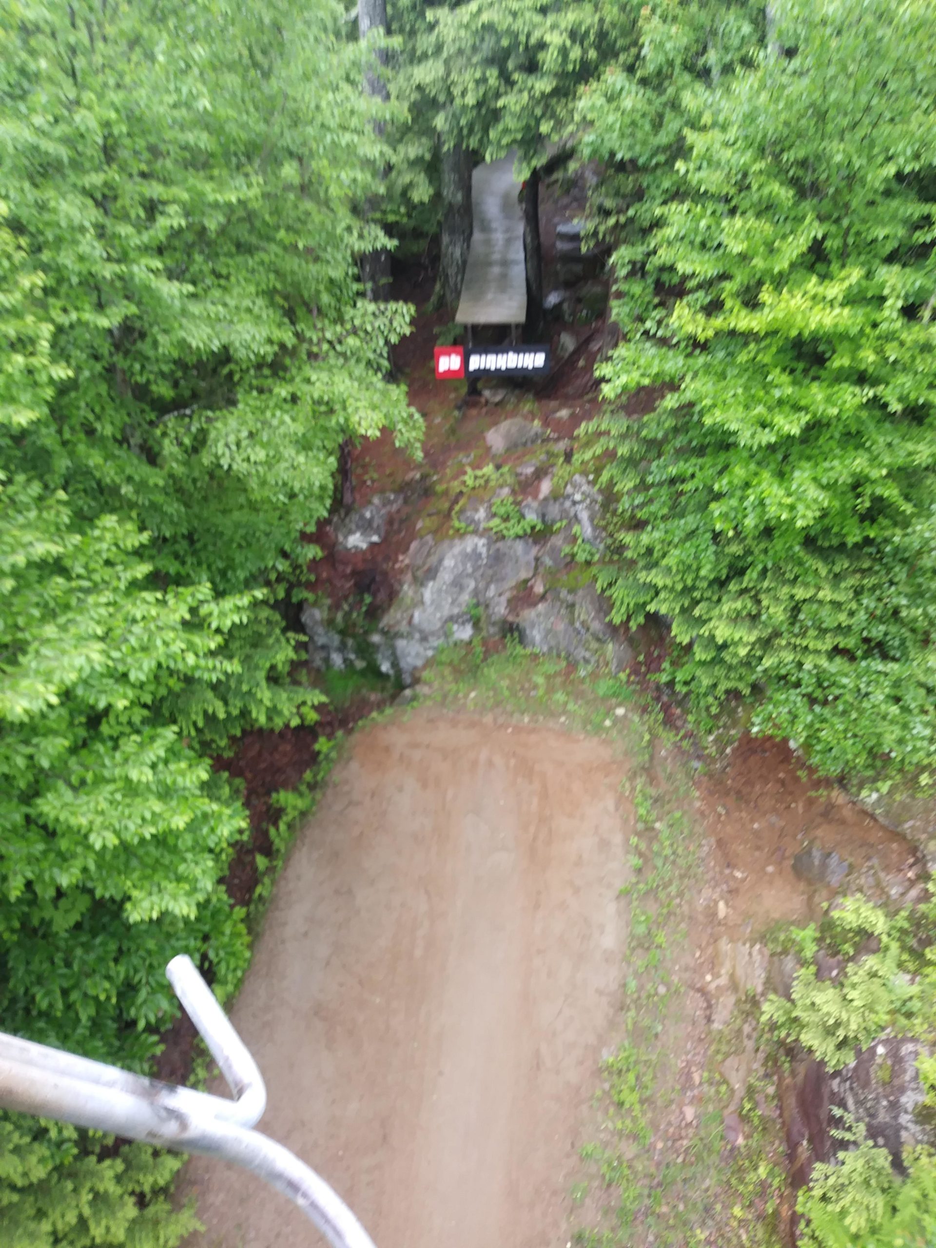 Aerial view of a dirt path leading through a lush green forest, with a wooden ramp extending over a rocky area. A sign is visible near the ramp, partially obscured by foliage. The scene is set in a natural environment, showcasing the rich greenery and earthy tones. Highland Mountain Bike Park mountain bike trail.