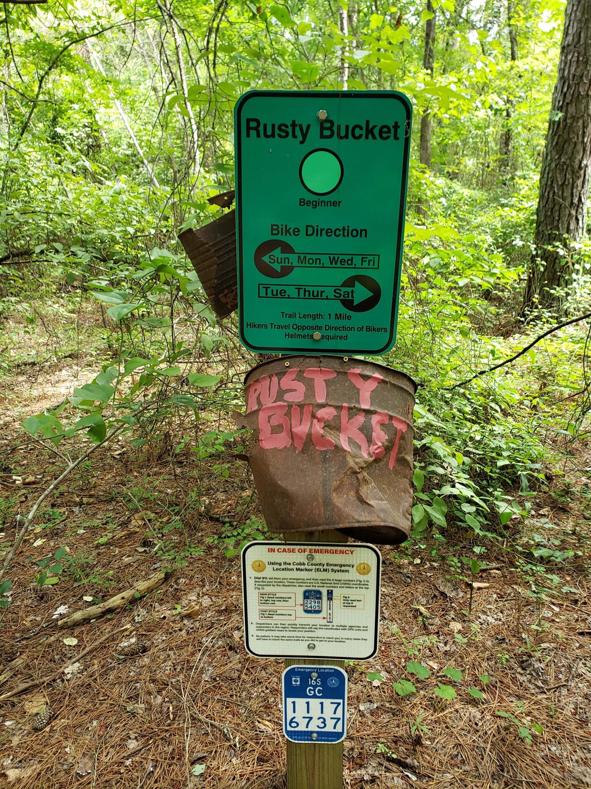 A green trail sign for the "Rusty Bucket" bike trail, indicating it is a beginner level route. The sign displays information about bike directions for specific days of the week and notes a trail length of one mile. Below the main sign, there is an emergency location marker with a blue background, providing instructions for emergencies. The trail area is surrounded by lush green trees and underbrush. Rusty Bucket Loop mountain bike trail.