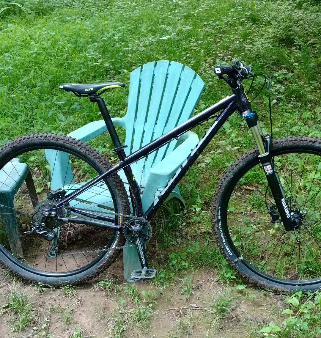 A mountain bike resting beside a teal Adirondack chair on a grassy area. The bike features a dark frame, thick tires with mud, and has a visible gear system. The background includes green foliage and natural vegetation. Great Bear mountain bike trail.