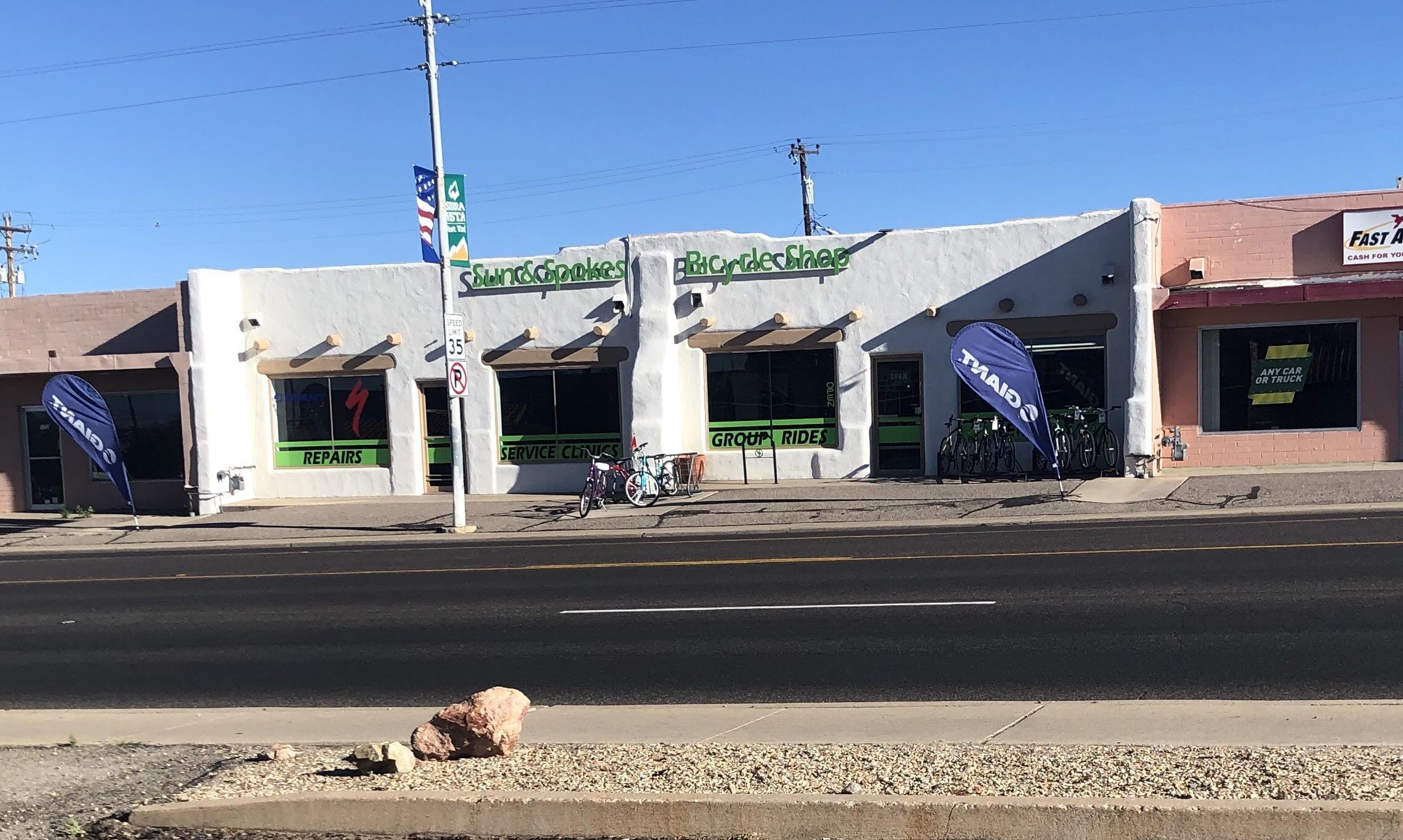 Image of a bicycle shop with a white adobe-style exterior featuring large windows. The storefront displays signs for "Repairs," "Service Clinics," and "Group Rides" in green lettering. Bikes are visible outside the shop, and banners promoting biking can be seen. A pink building is adjacent to the shop, displaying a sign for cash services. The scene is set under a clear blue sky.
