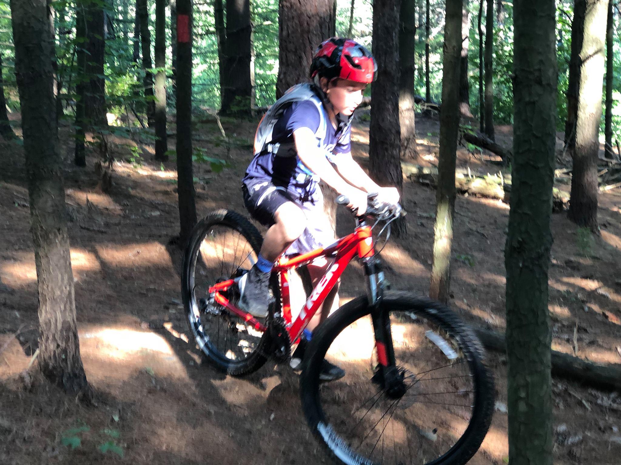 A young cyclist riding a red mountain bike on a dirt trail through a forest. Sunlight filters through the trees, casting soft shadows on the ground. The cyclist is wearing a helmet and athletic gear, focused on navigating the rough terrain. Bavington mountain bike trail.
