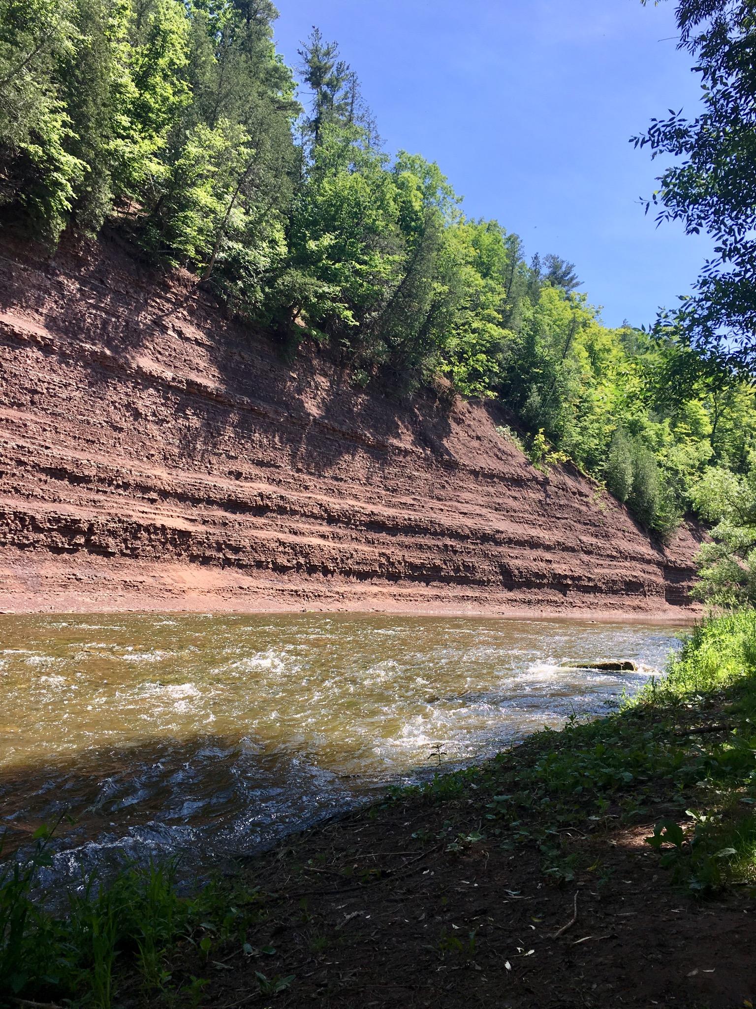 A serene river flowing alongside a steep, layered reddish-brown cliff, with dense greenery on top and beside the water under a clear blue sky. 16 Mile Creek - Lions Valley Park mountain bike trail.