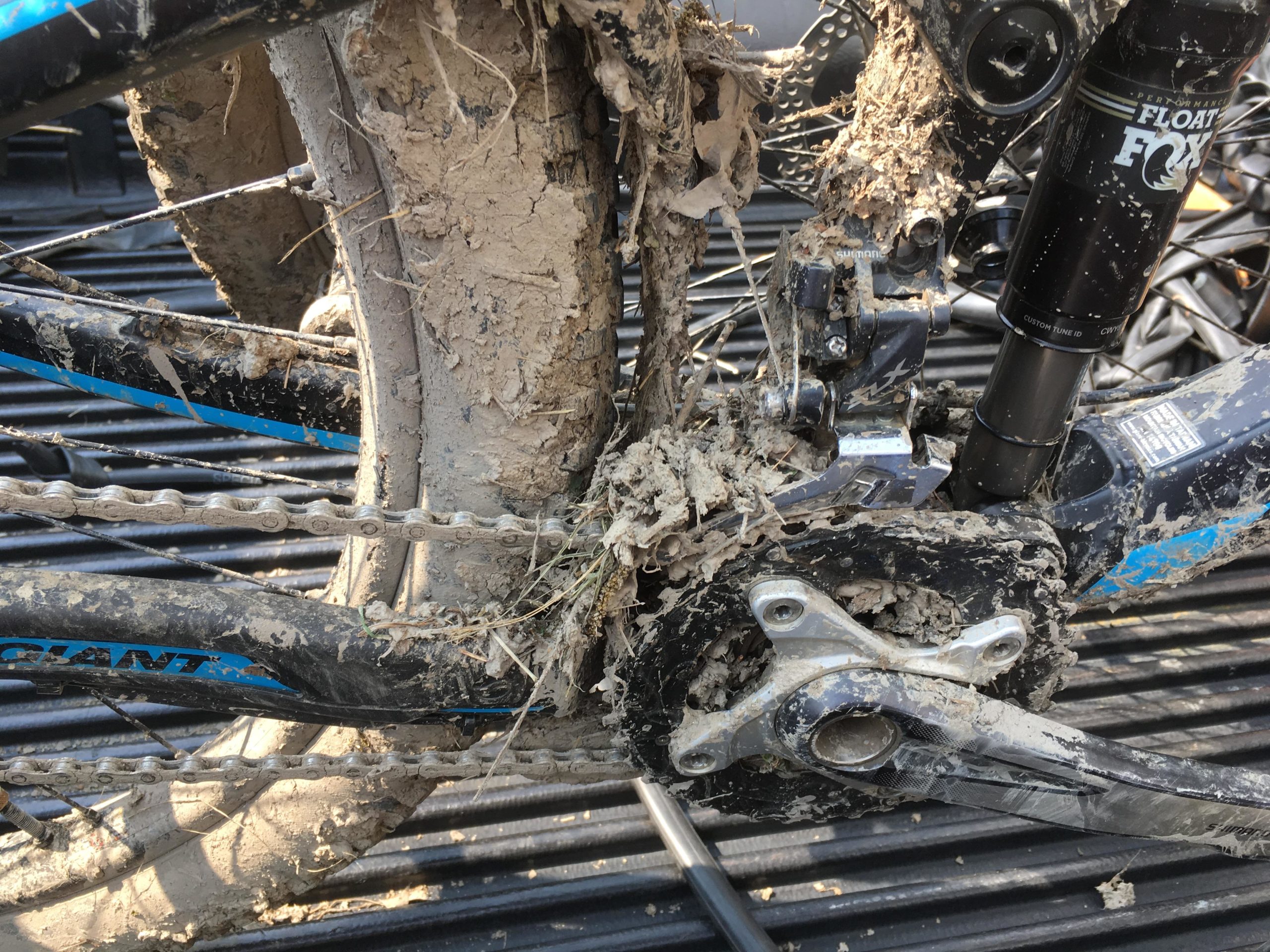 Close-up image of a mountain bike's rear drivetrain, heavily caked with mud and debris. The chain, gears, and frame are visibly dirty, showcasing signs of recent off-road use. The background shows a textured, possibly truck bed surface. Stewart State Forest mountain bike trail.