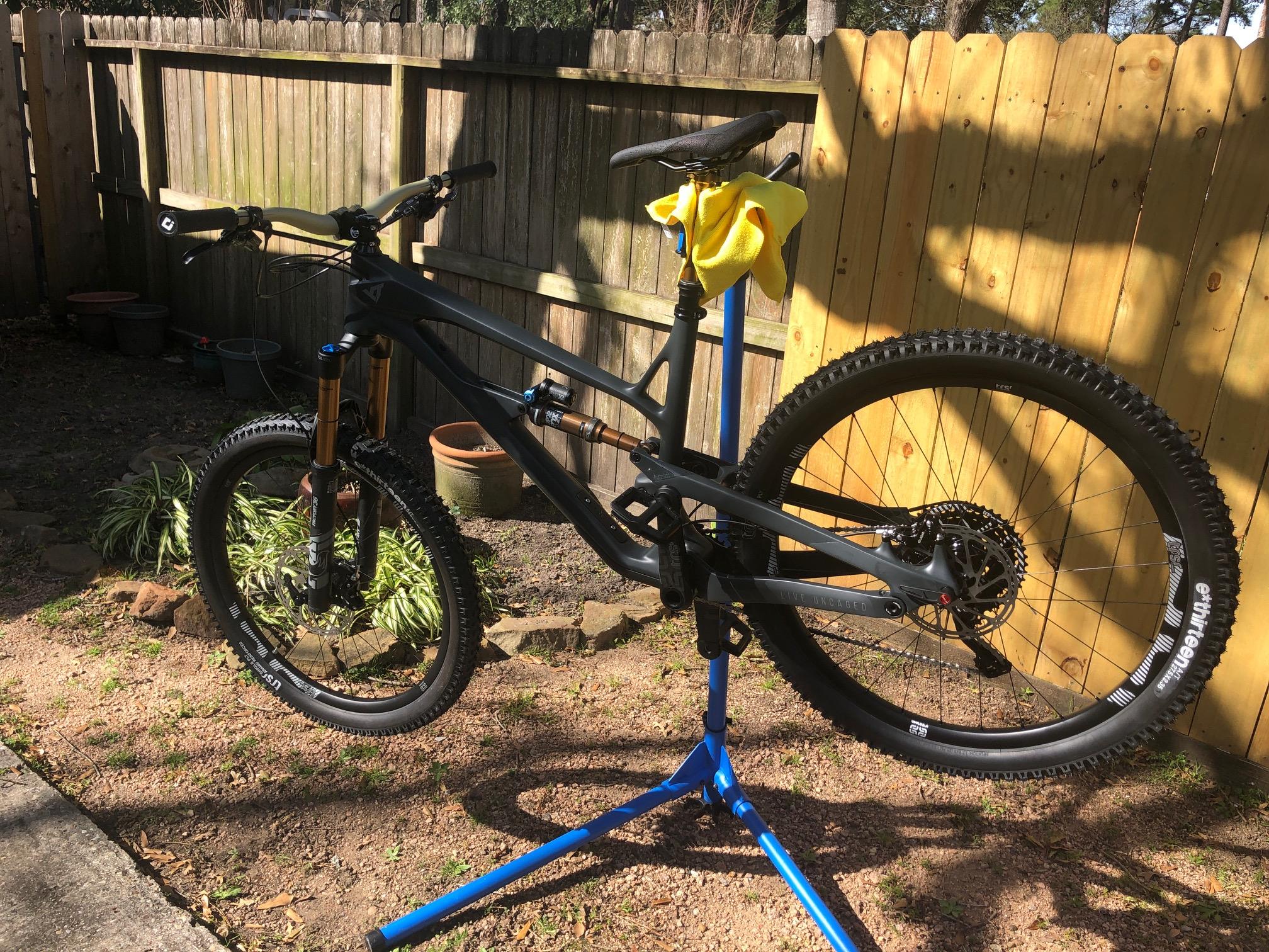 YT Industries YT Jeffsy CF: A mountain bike on a blue repair stand, positioned outdoors. The bike features a black frame with gold accents, thick tires, and front suspension. A yellow cloth is draped over the seat. In the background, there is a wooden fence and potted plants. Sunlight is casting shadows in the scene.