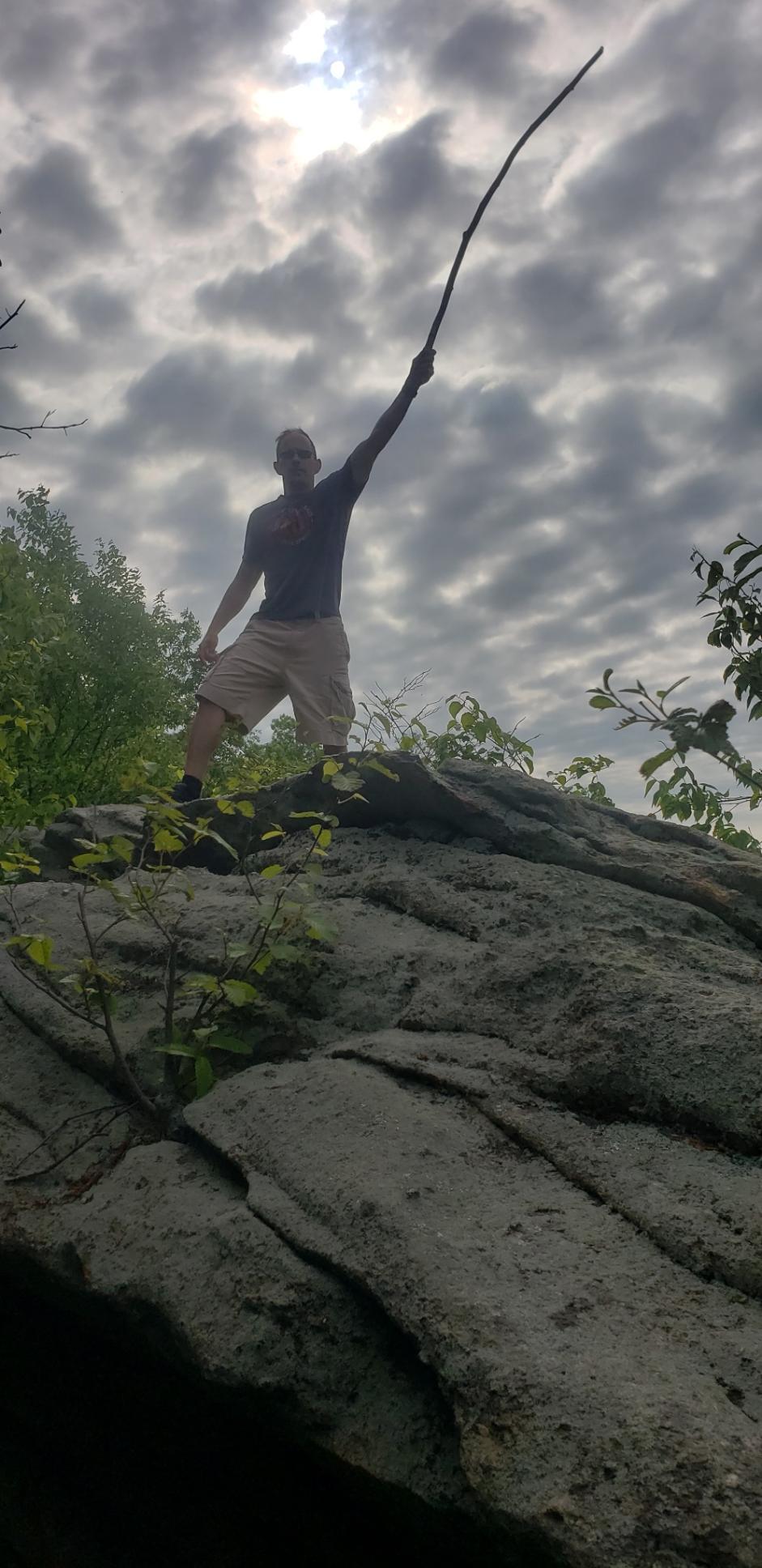 Trek Fuel EX 7 29: A person standing on a large rock with one arm raised, holding a stick, against a backdrop of cloudy skies and greenery. The individual is wearing a t-shirt and shorts, projecting a sense of adventure or triumph.