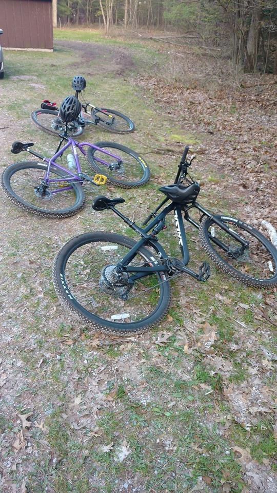 Salsa Spearfish: Three mountain bikes are resting on a dirt path covered with fallen leaves. A brown shed is visible in the background, surrounded by trees. Two of the bikes have helmets placed on them.