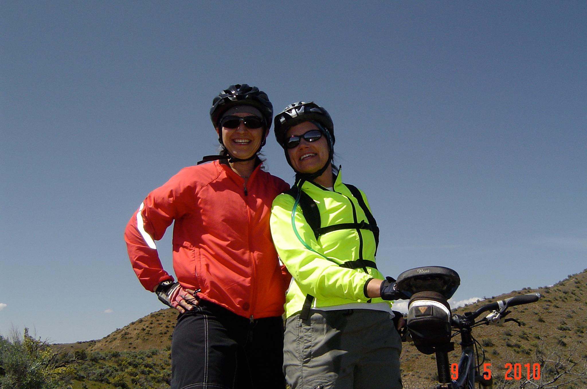 Yeti ASR Five Carbon: Two women, wearing cycling helmets and sunglasses, smile together in a sunny outdoor setting. One is dressed in a bright red jacket and the other in a neon yellow jacket, both with biking gear. They stand beside a bicycle in a mountainous landscape under a clear blue sky.