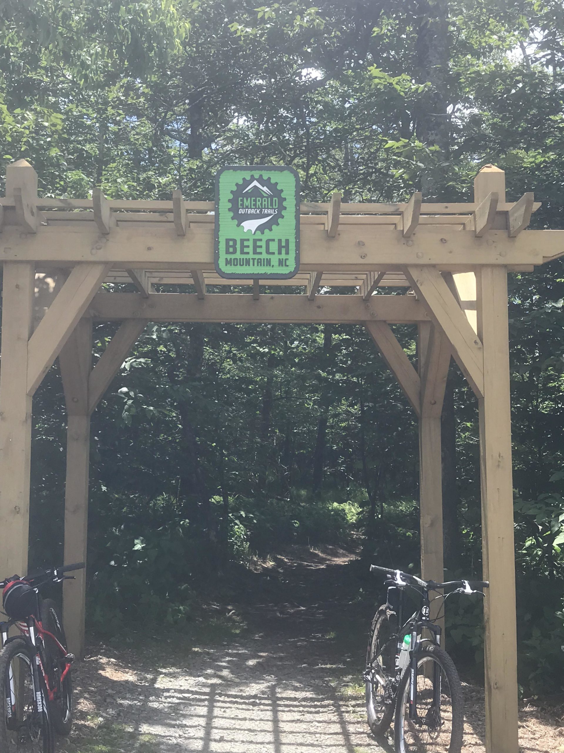 Archway sign for Emerald Outback Trails at Beech Mountain, NC, with two mountain bicycles parked on either side of a gravel path leading into a wooded area. The scene is surrounded by lush greenery and dappled sunlight filtering through the trees. Emerald Outback mountain bike trail.