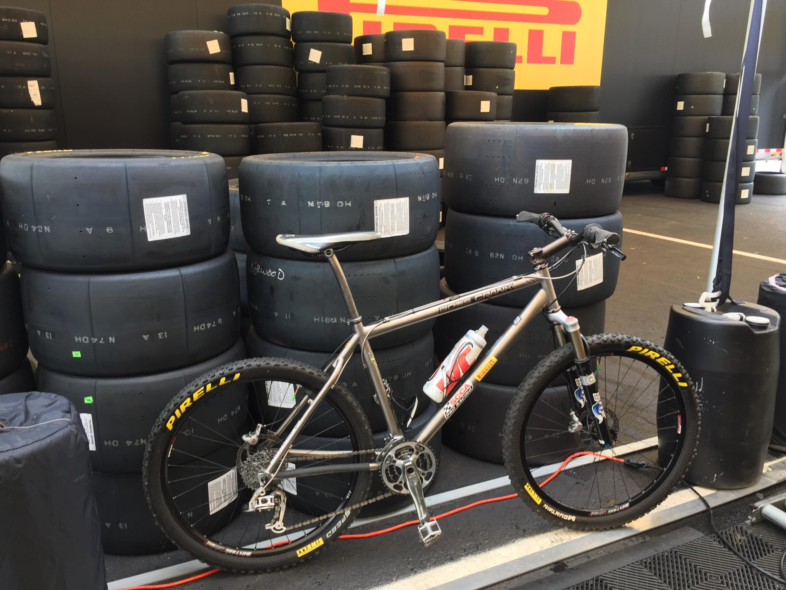 Gary Fisher Paragon: A mountain bike with a metallic frame is positioned next to a stack of racing tires. The tires are black and prominently display the Pirelli logo, with labels visible on several of them. The background features a dark-colored wall with more tires stacked up, creating an industrial racing environment.