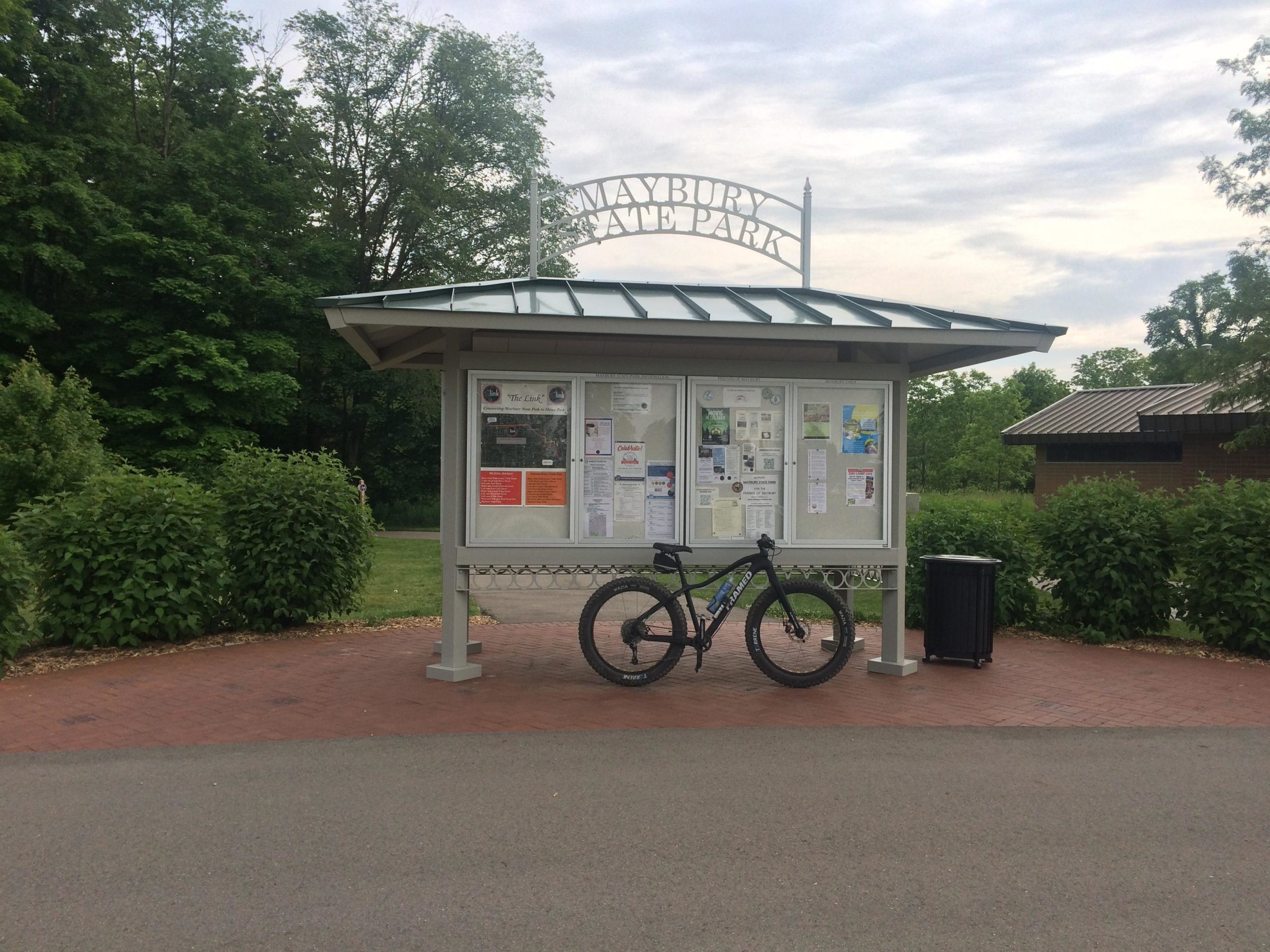Information board at Maybury State Park with a bicycle leaning against it, surrounded by green foliage and walking paths. A building is visible in the background under a cloudy sky. Maybury mountain bike trail.