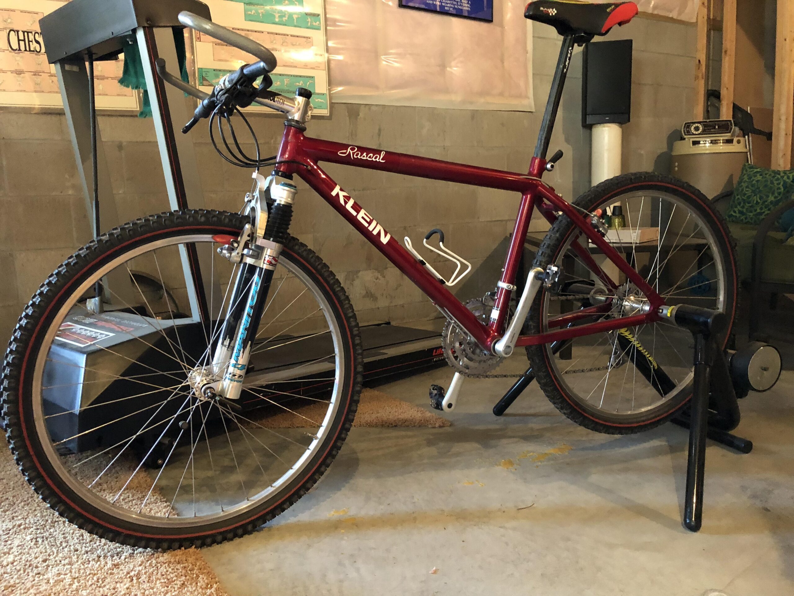 Gary Fisher Paragon: A maroon Klein mountain bike with front suspension is positioned on a bike stand in a garage setting, alongside a treadmill and other fitness equipment. The bike features black tires with red accents and a comfortable seat. The walls are made of concrete blocks and the floor is covered with a light carpet.