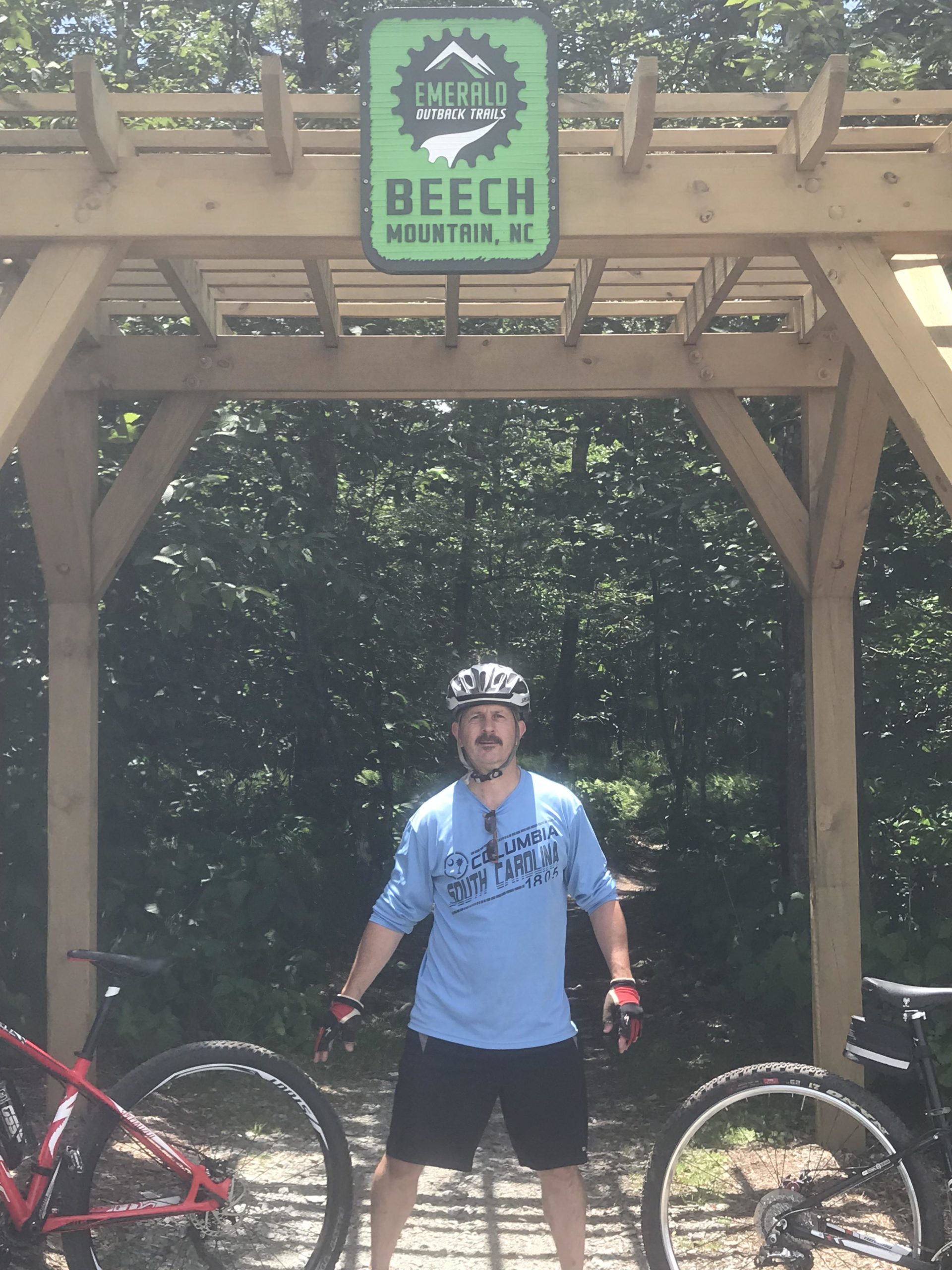 A mountain biker stands in front of a wooden archway sign that reads "Emerald Outback Trails, Beech Mountain, NC." The sign is surrounded by lush greenery, and two bikes are positioned beside the cyclist, who is wearing a helmet and sport attire. The scene suggests an outdoor adventure in a mountainous area. Emerald Outback mountain bike trail.