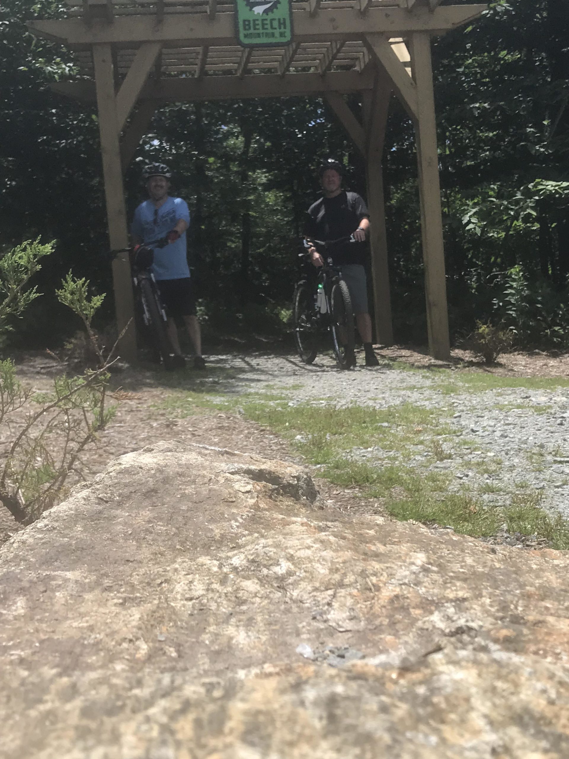Two men stand with their bicycles under a wooden archway marked with a sign that reads "Beech Mountain, NC." The background is lush with greenery, suggesting a forested area. A large rock is visible in the foreground, and the ground is a mix of dirt and gravel. The scene is bright and sunny, indicating a clear day for outdoor activities. Emerald Outback mountain bike trail.