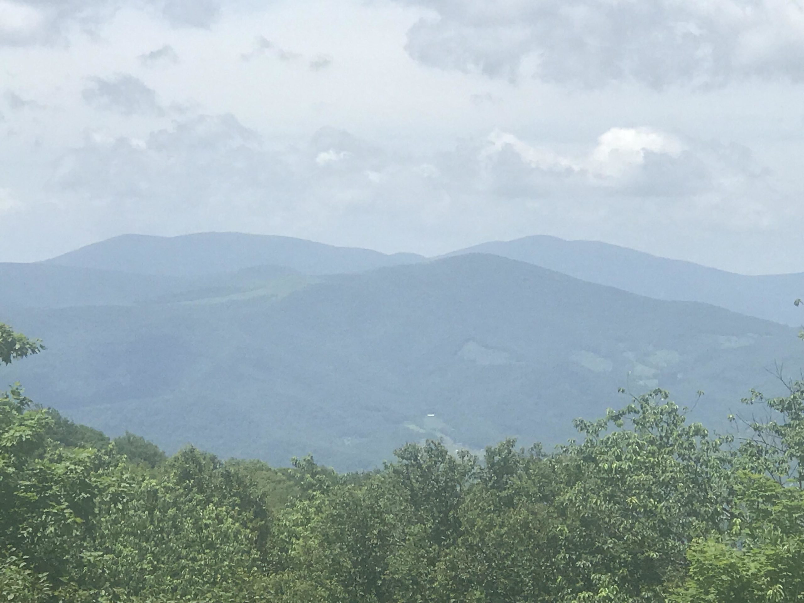 A scenic view of rolling mountains under a cloudy sky, with varying shades of green from trees in the foreground and the distant blue-gray hues of the mountains. Emerald Outback mountain bike trail.
