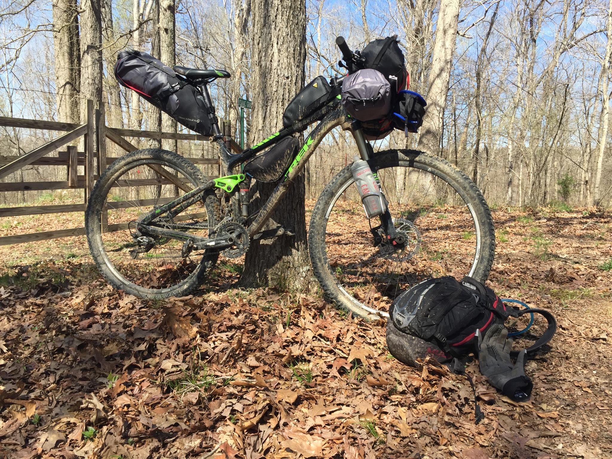Trek Fuel EX 7 29: A mountain bike with attached saddle and frame bags is leaning against a tree in a forested area, surrounded by fallen leaves. A pair of gloves and a hydration pack are on the ground nearby, suggesting a biking adventure. The scene depicts a clear day with trees in the background and a wooden fence in the distance.