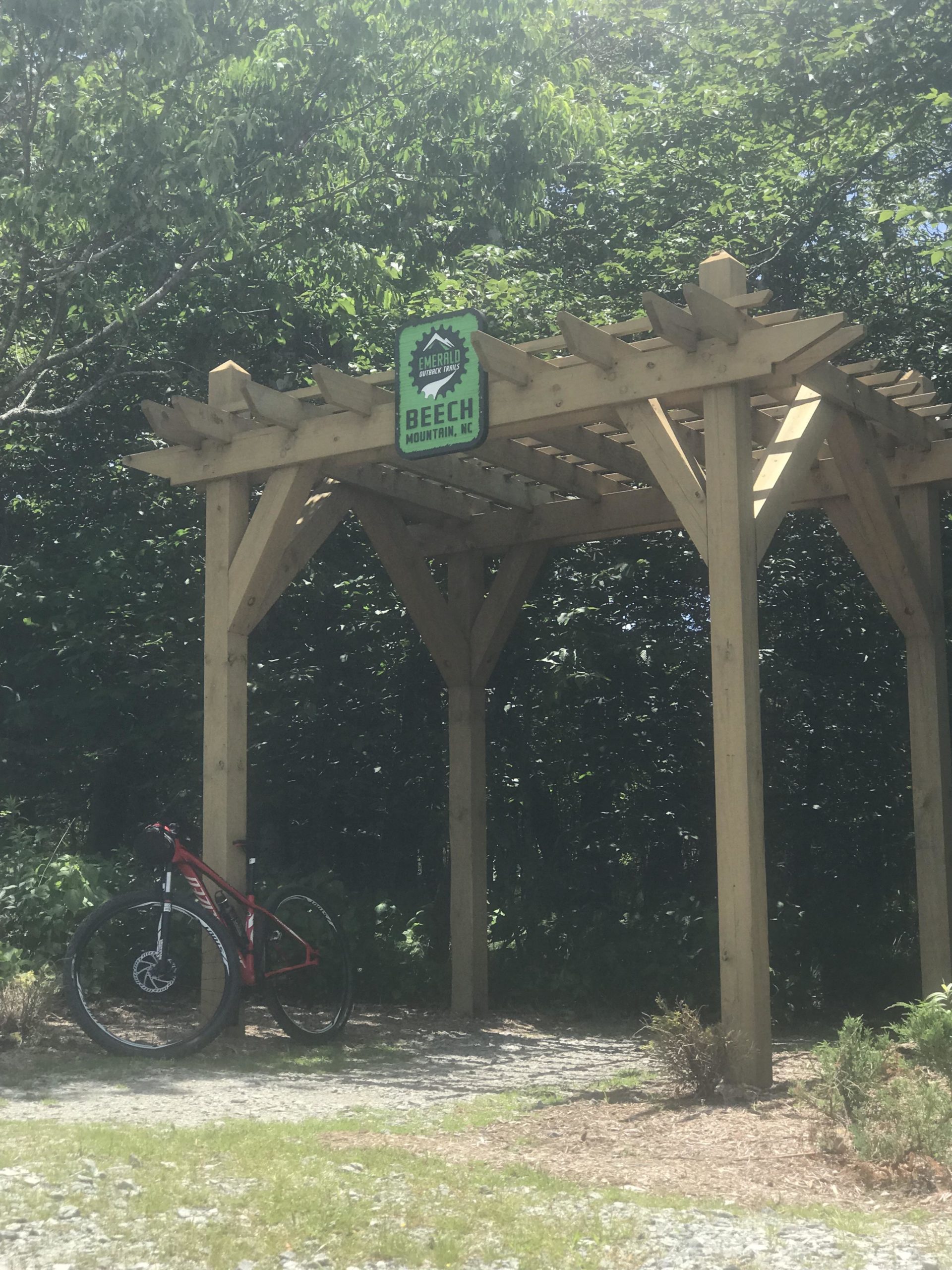 Sign for Beech Mountain, NC, displayed on a wooden structure in a forested area. A red bicycle is leaning against the structure, surrounded by greenery and gravel pathways. Emerald Outback mountain bike trail.