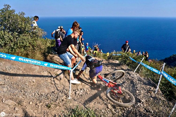 A mountain biker falls near the edge of a steep, rocky trail while others watch from above, with a blue race barrier prominently displayed. The ocean can be seen in the background under a clear sky, and the scene captures the excitement and intensity of a mountain biking event.