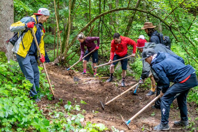 A group of six individuals, dressed in outdoor gear, are working together in a lush, green forest area. They are using tools such as rakes and shovels to maintain or create a trail in the dirt. The scene showcases teamwork and outdoor activity, surrounded by trees and vibrant greenery.