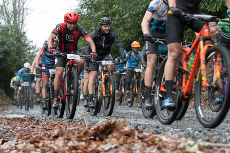 A group of mountain bikers participating in a race on a gravel trail during rainy weather, surrounded by greenery and leaves on the ground. The cyclists are wearing colorful cycling jerseys and helmets, with a focus on a rider in the foreground wearing a red helmet and jersey.