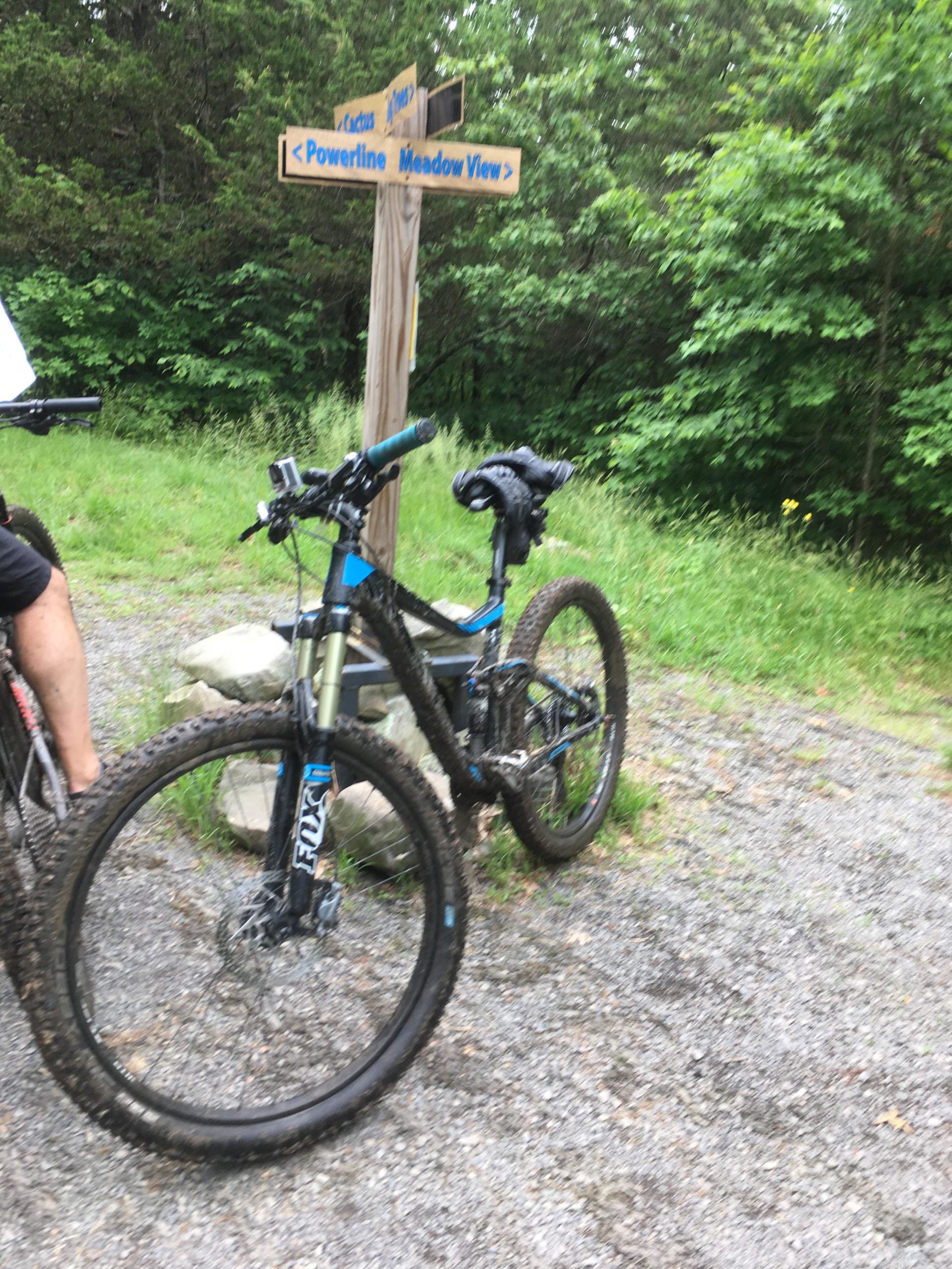 A mountain bike with a muddy frame and tire rests next to a wooden trail sign indicating directions to "Powerline" and "Meadow View." Surrounding greenery is vibrant, and gravel paths are visible, suggesting an outdoor biking location. Stewart State Forest mountain bike trail.