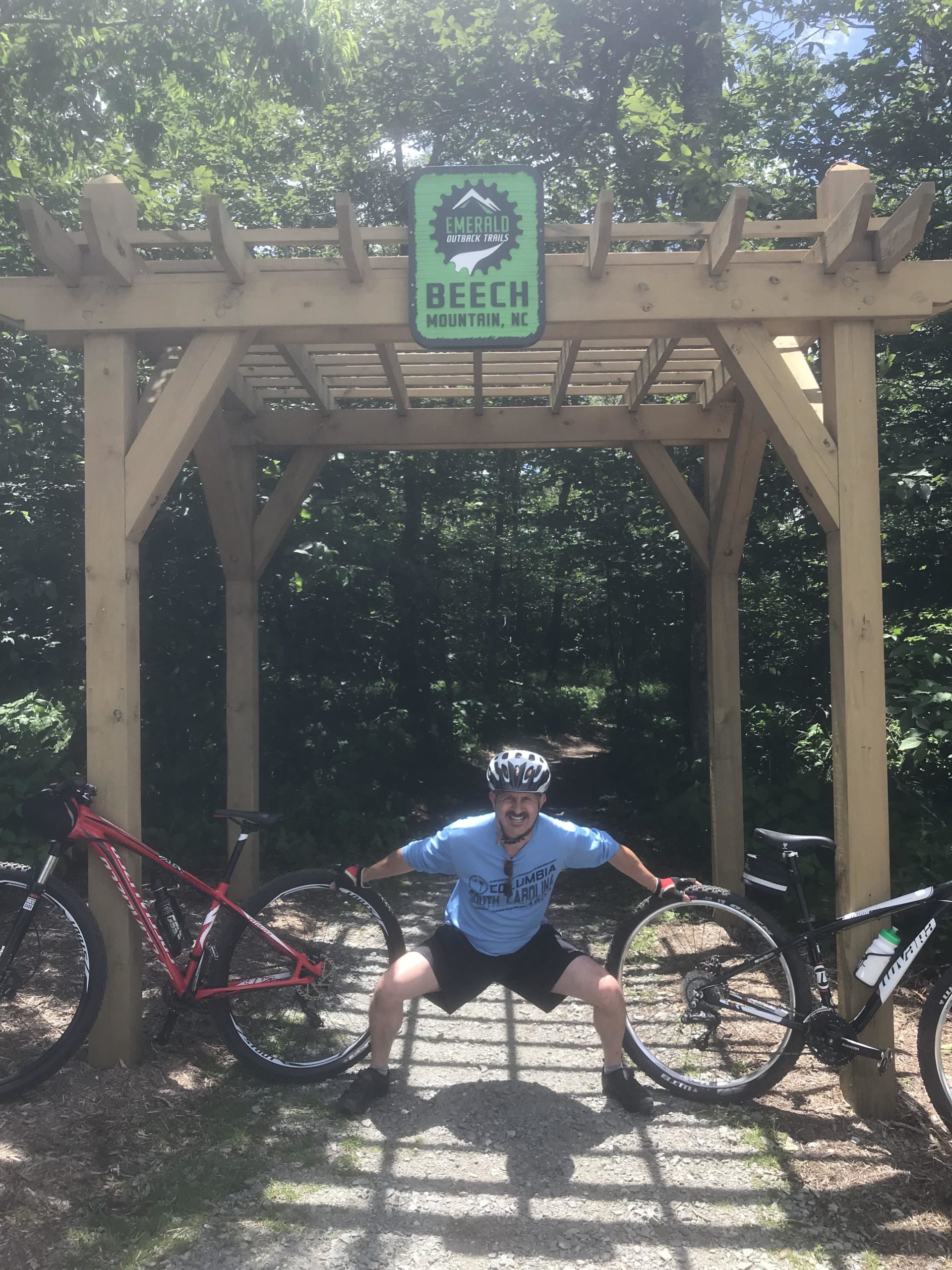 A man in a blue shirt and helmet strikes a playful pose while holding two bicycle wheels at the entrance of the Emerald Outback Trails at Beech Mountain, NC. Two mountain bikes are leaning against a wooden archway with a sign indicating the trail's name. Sunlight filters through the trees above. Emerald Outback mountain bike trail.