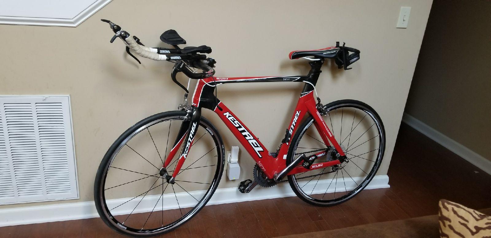 Gary Fisher Paragon: A red Kestrel road bicycle leaning against a wall in a room with wooden flooring. The bike features aero handlebars and a sleek design, with visible components including gears and tires. An air vent is located on the wall beside the bike, and a small wall plug can be seen behind it. The interior has a neutral wall color and part of a patterned chair is visible in the foreground.