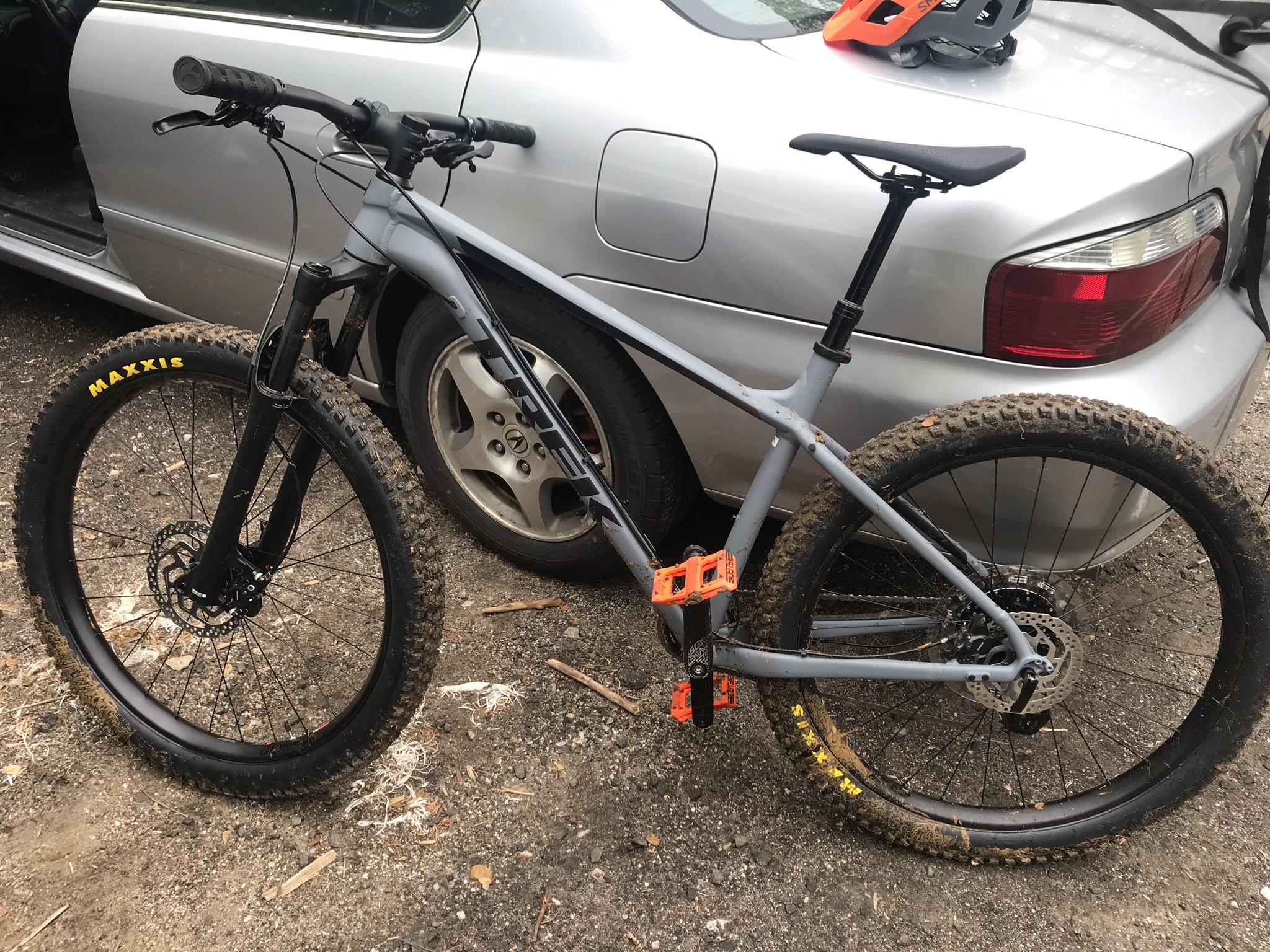 Trek Roscoe 8: A gray mountain bike with black tires and orange pedals parked next to a silver car, with a helmet on the car's roof. The bike's tires are muddy, indicating recent use on trails.