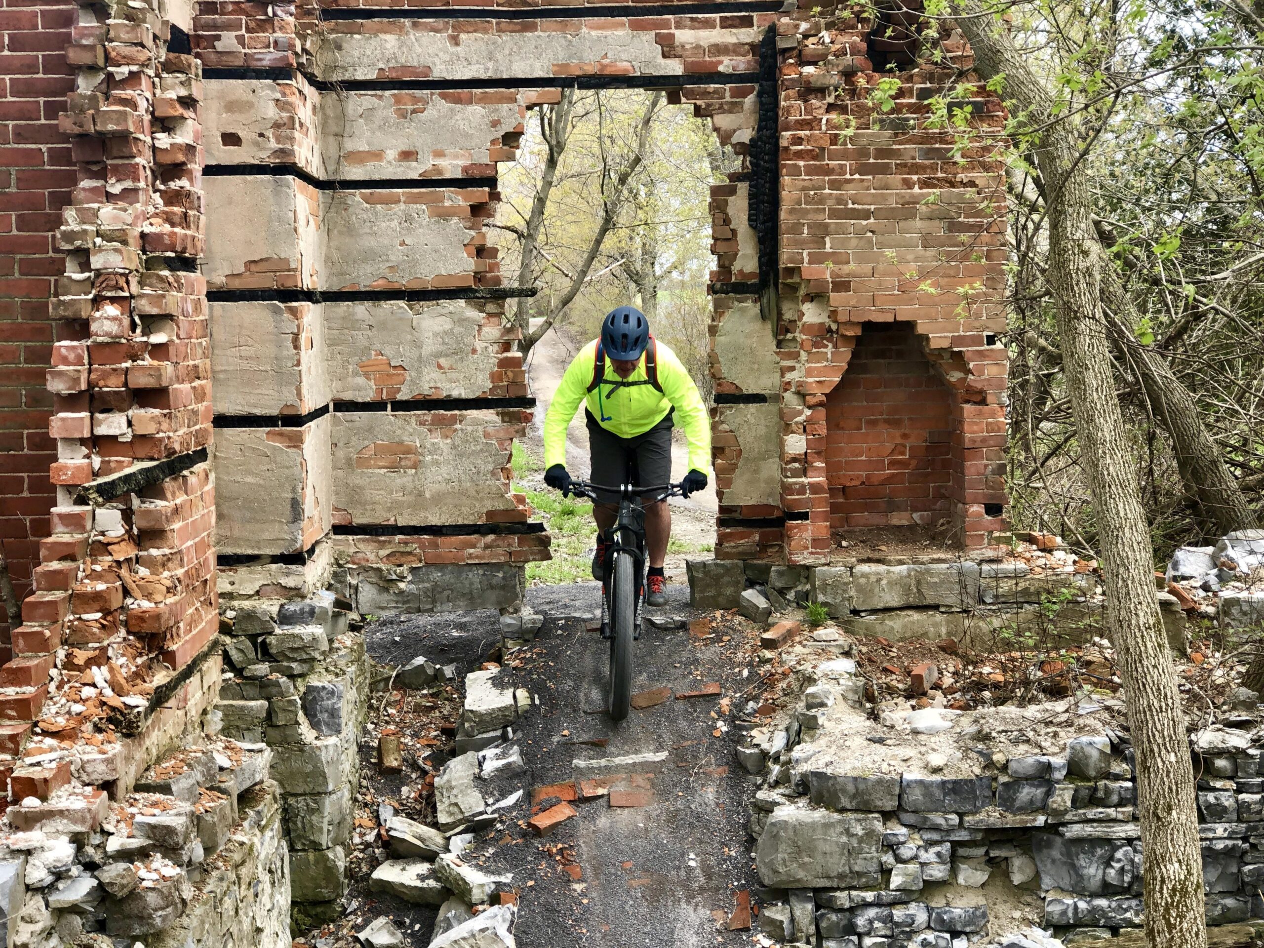 Trek Trek Stache 9.7: A person wearing a bright yellow jacket and a helmet rides a mountain bike across the remnants of a brick structure in a natural setting. The scene features crumbling walls, exposed bricks, and overgrown vegetation, creating a contrast between the man-made ruins and the surrounding greenery.