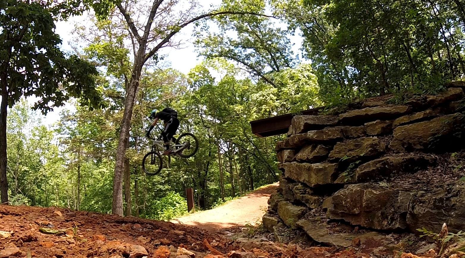 A cyclist performing a jump on a mountain bike over a dirt path, with a rocky outcrop on the right and trees in the background. The scene captures a moment of action amidst a natural wooded environment. Coler Mountain Bike Preserve mountain bike trail.